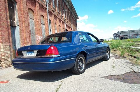 2001 Ford Crown Victoria - A blue 2001 Ford Crown Victoria is parked on a cracked asphalt surface near an old brick building under a bright blue sky.