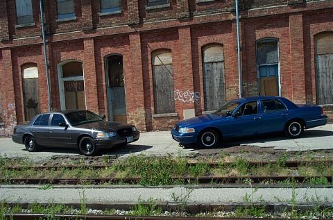 2001 Ford Crown Victoria - Two Ford Crown Victorias are parked side-by-side in front of an old brick building.
