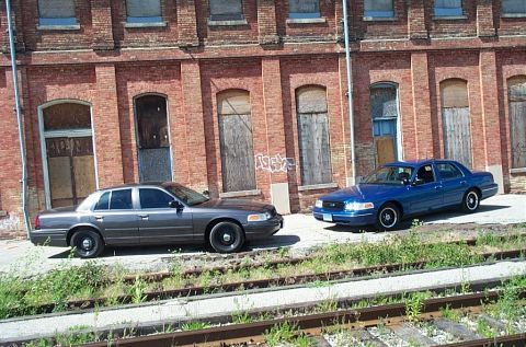 2001 Ford Crown Victoria - Two Ford Crown Victorias are parked side-by-side on gravel near railroad tracks, with a brick building behind them.