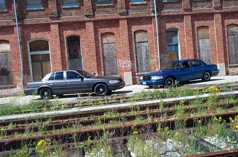 2001 Ford Crown Victoria - Two Ford Crown Victoria police cars are parked on train tracks in front of a brick building.