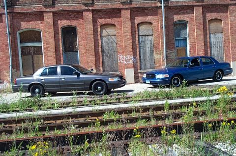 2001 Ford Crown Victoria - Two Ford Crown Victorias are parked side-by-side in front of an old brick building.