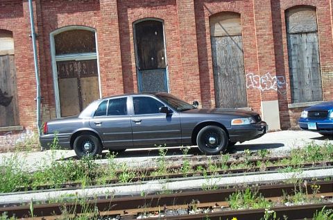 2001 Ford Crown Victoria - A dark gray Ford Crown Victoria is parked beside an old brick building with boarded-up windows.