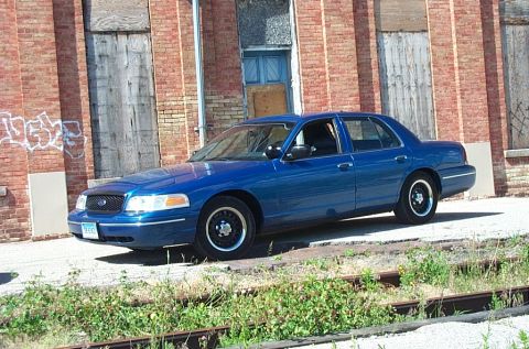 2001 Ford Crown Victoria - A blue 2001 Ford Crown Victoria sedan is parked outdoors in front of a brick building with boarded-up windows.