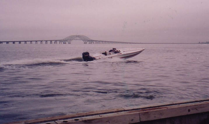 White outboard speedboat running on gray bay water near a long causeway bridge