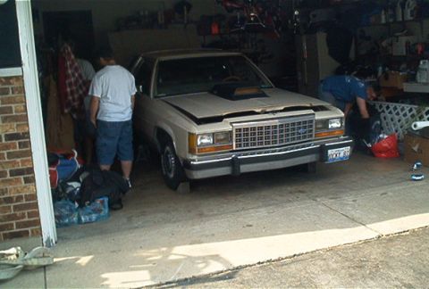 1985 Ford LTD Crown Victoria - Tan two-tone Ford LTD Crown Victoria sedan backed into a home garage with the hood off and the engine bay open, photographed from the front…