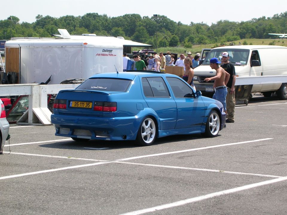 Blue sedan with custom body kit and aftermarket wheels parked in a lot. White trailer and van in background