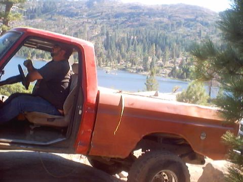 1976 Ford F-100 - A man drives a red pickup truck with a lake and mountains in the background.