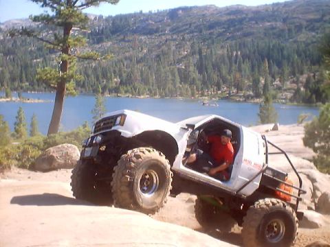 1976 Ford F-100 - A modified white truck navigates a challenging off-road trail with a scenic lake in the background.