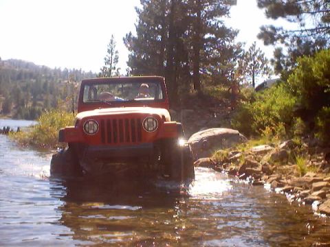 1976 Ford F-100 - A red Jeep navigates through a rocky stream, with trees and a lake visible in the distance.