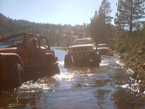1976 Ford F-100 - Several off-road vehicles navigate a water crossing on a sunny day, with a wooded hillside in the distance.