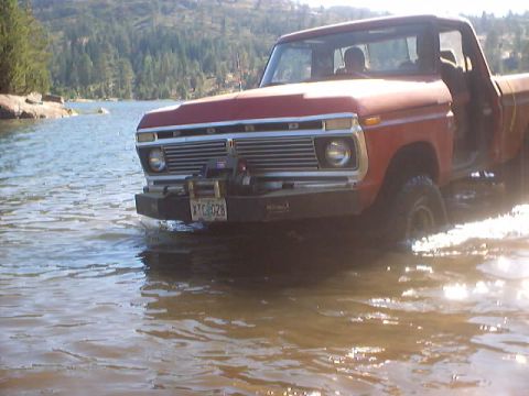 1976 Ford F-100 - A red pickup truck with a winch on the front bumper drives through shallow water, with trees and a lake in the background.