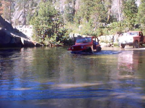 1976 Ford F-100 - Two off-road vehicles navigate a river crossing surrounded by trees and rocky terrain.