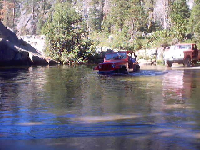 Red Jeep Wrangler and a white and orange truck crossing a river in a wooded area