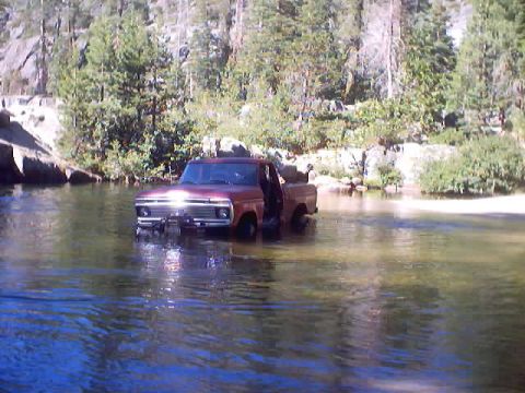 1976 Ford F-100 - A red pickup truck is partially submerged in a river, surrounded by trees and rocky terrain.