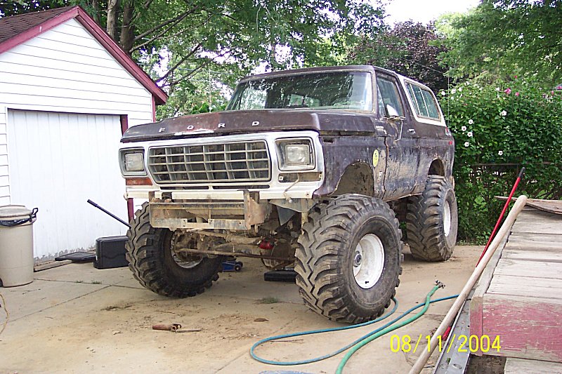 Lifted dark-brown Ford Bronco with big mud tires on white steel wheels, parked by a white shed