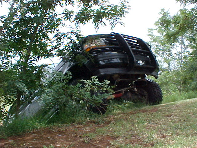 Dark lifted Ford with brush guard climbing a grassy bank, front axle visible