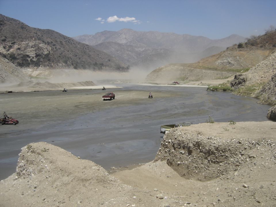 Vehicles kicking up dust crossing a wide gray riverbed in an arid canyon ringed by mountains
