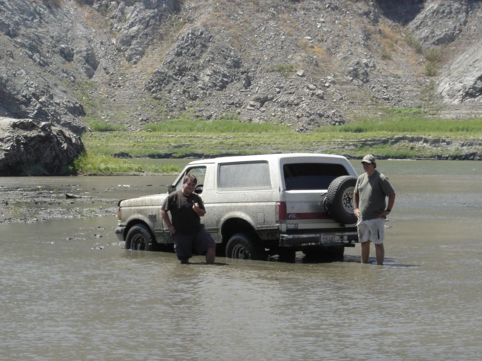 Muddy tan Ford Bronco stuck in shallow river water with two men standing beside it