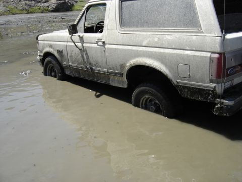1988 Ford Bronco - A white Ford Bronco is stuck in a muddy puddle, with its tires submerged.