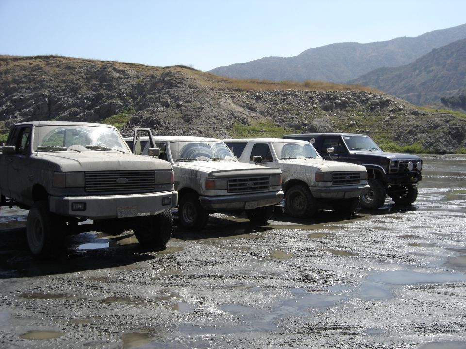 Row of four muddy lifted Ford trucks and Broncos parked on wet mud with hills in background