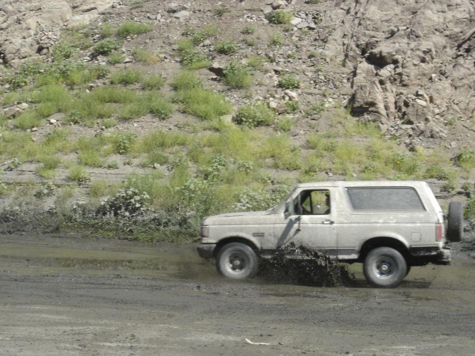 Muddy tan Ford Bronco splashing through mud in side view, rocky hillside behind