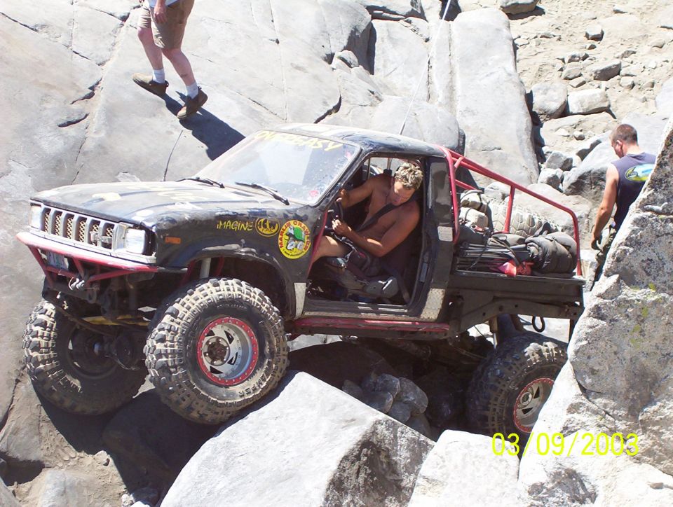 Off-road truck with large tires climbing over rocks. Driver is shirtless