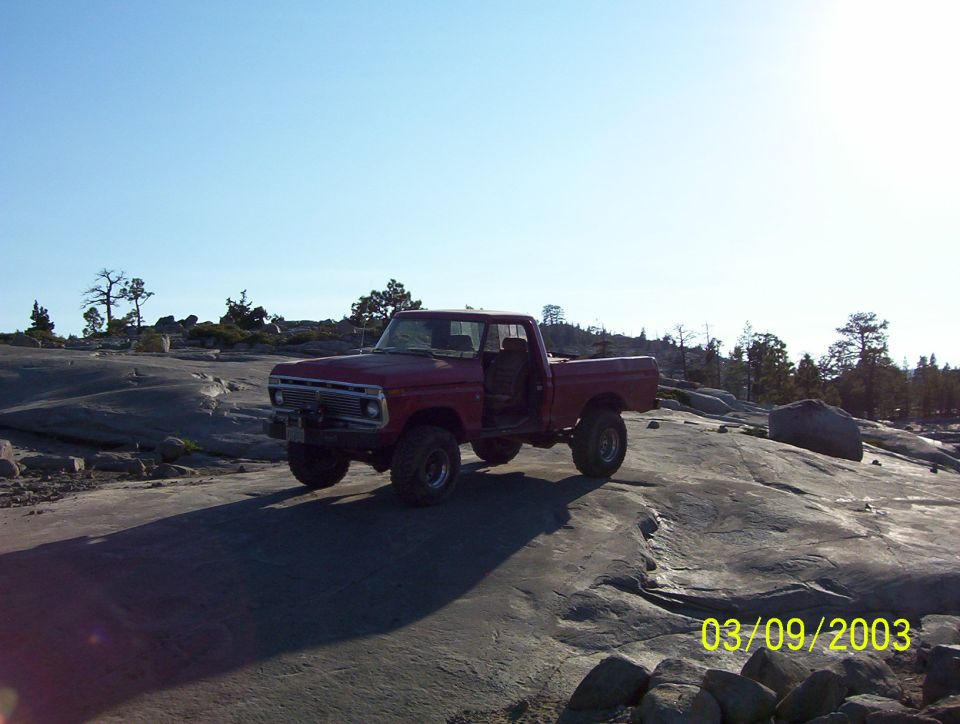 Red 1975 Ford F-100 pickup truck with doors open, parked on a rocky landscape under a clear blue sky