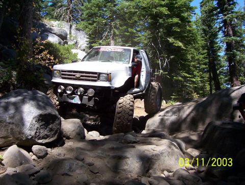 1976 Ford F-100 - This modified truck is shown tackling a challenging off-road course with large boulders and trees surrounding it.