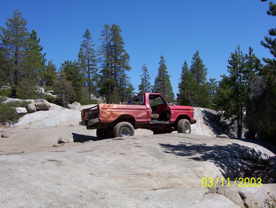 Faded red doorless square-body Ford pickup crawling over granite slab in pine forest, 2003 date stamp