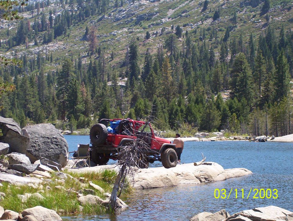 Red topless Jeep Wrangler with spare tire parked on granite lakeshore in a pine-covered mountain basin