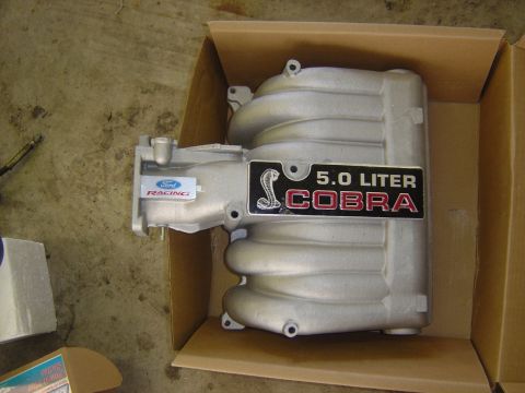 1993 Ford Bronco - A Ford Racing GT40 upper intake manifold sitting in its open cardboard box on a concrete floor, cast aluminum with a black '5.0 LITER COBRA…