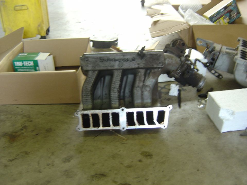 Grimy stock upper intake manifold removed and set on a shop floor beside parts boxes