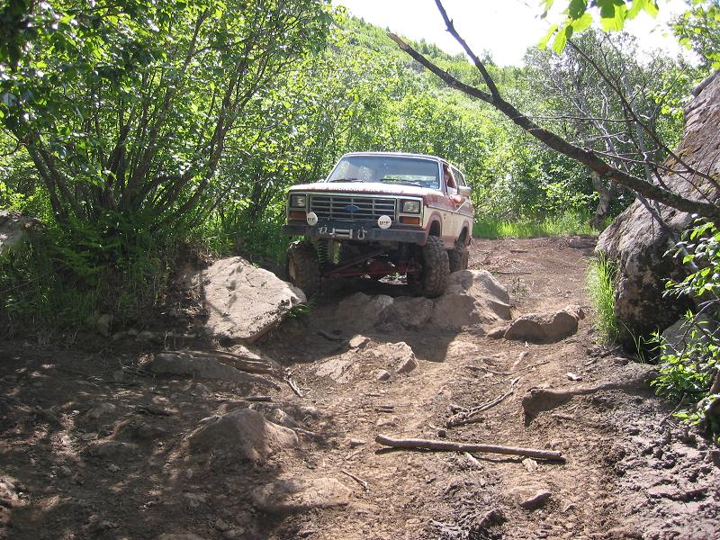 Red and white lifted Ford Bronco crawling down a rocky trail flanked by green brush