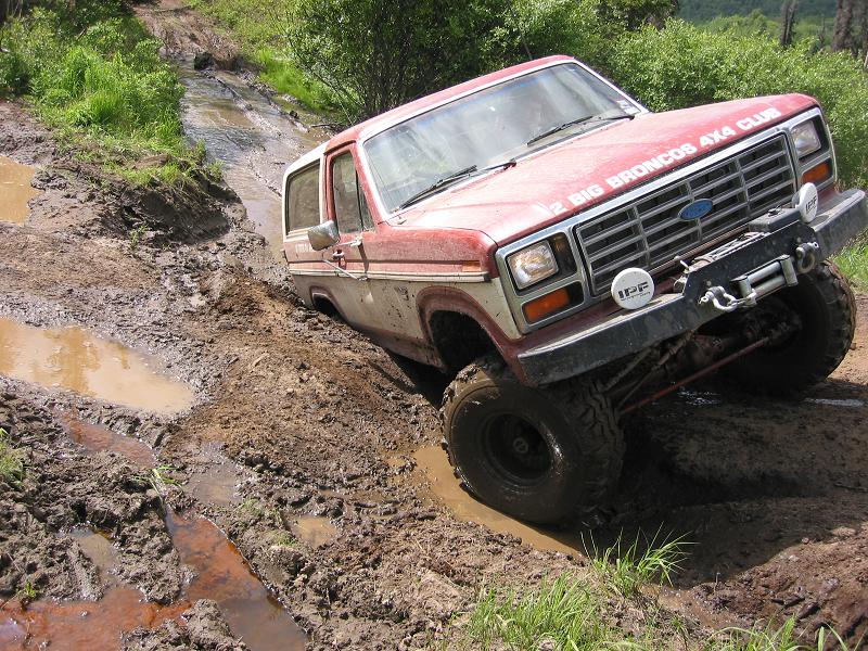 Faded red Ford Bronco 4x4 crawling deep mud ruts, front three-quarter view