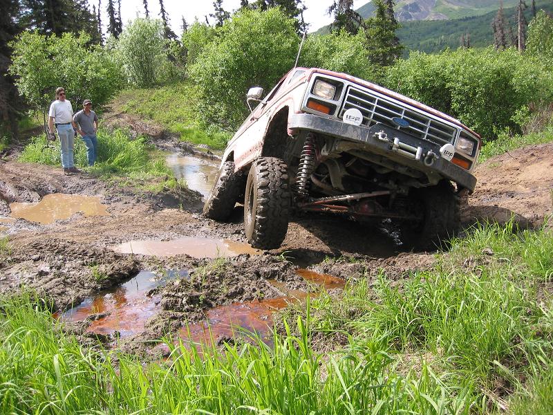 Red and white lifted Ford Bronco flexing over a muddy trail rut with winch and IPF lights