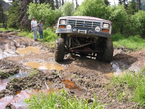 1985 Ford Bronco - A lifted Ford Bronco navigates a challenging muddy trail with two onlookers observing the off-road adventure.