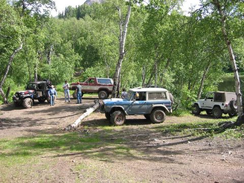 1985 Ford Bronco - A blue Ford Bronco is parked in a clearing surrounded by trees, with several other off-road vehicles nearby.