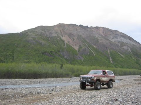 1985 Ford Bronco - 1985 Ford Bronco driving through a rocky riverbed with a mountain range behind it.