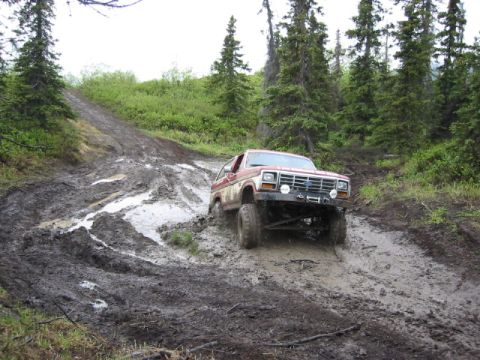 1985 Ford Bronco - A red and white Ford Bronco navigates a challenging muddy terrain on an off-road trail.