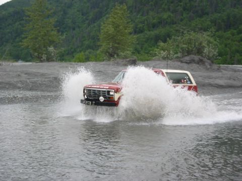 1985 Ford Bronco - SUV navigates a water crossing, kicking up a large spray.