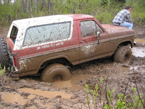 1985 Ford Bronco - A red and white 1985 Ford Bronco is deeply stuck in a muddy mire, with a person sitting on its hood.