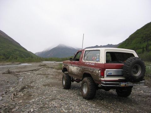1985 Ford Bronco - 1985 Ford Bronco sits on a rocky, muddy terrain with a river and mountains behind it.