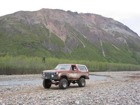 1985 Ford Bronco - A lifted Ford Bronco sits on a rocky riverbed, with a large mountain range dominating the background.