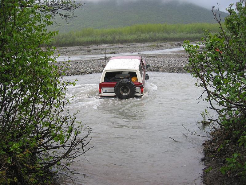 Red and white Ford Bronco with rear-mounted spare crossing a silty river, water at rocker height