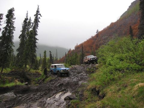 1985 Ford Bronco - Two SUVs tackle a challenging, muddy off-road path in a remote, mountainous area.