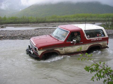 1985 Ford Bronco - A red and white 1985 Ford Bronco navigates a river, with visible text on its rear window indicating it's part of a 4x4 club.