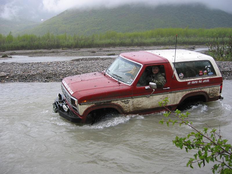 Red-and-white Ford Bronco crossing a silty river, winch bumper, front 3/4.
