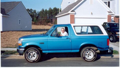 1995 Ford Bronco - A blue 1995 Ford Bronco is parked on the street in front of a residential building, with a man visible inside.