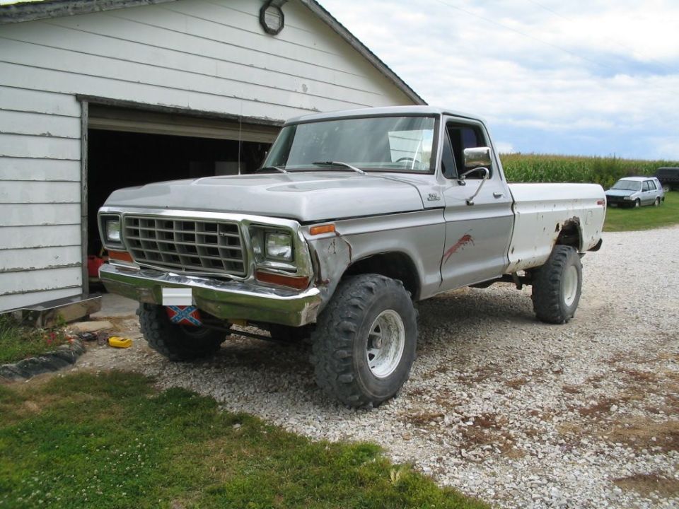 Silver 1979 Ford F-150 pickup truck with lifted suspension and large tires, parked in front of a garage