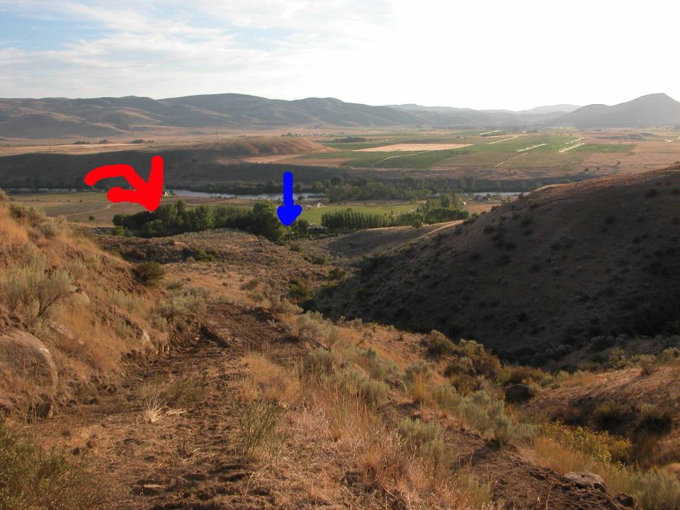 Dirt trail overlooking a river valley with hills, marked by red and blue arrows